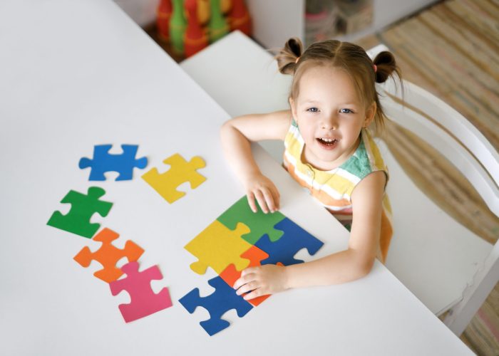 A laughing sweet girl is sitting at a table and assembling a puzzle of colorful elements on autism Day. Logical thinking and development. The child looks up into the camera.